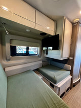 Inside an RV rental: a modern dinette area with sage green cushions, overhead storage, and a mounted TV.
