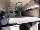 Cozy RV rental interior with a bed featuring chevron cushions, overhead cabinets, ceiling lights, and an AC unit.