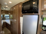 Modern RV rental interior: compact kitchen with microwave, fridge, and built-in storage leading to a cozy bed.