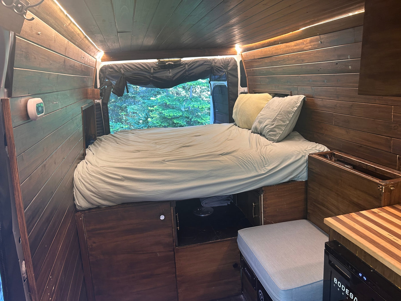 This RV rental's cozy interior features a comfortable bed, warm wooden paneling, ambient lighting, and a window showing lush trees. — photo 10