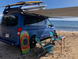 Beachside RV rental setup: a blue camper van with pop-top, awning, surfboard, chairs, and table by the ocean.
