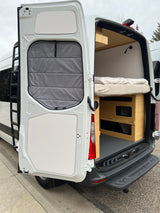 Rear view of an RV rental: white camper van with open doors, showcasing a cozy bed, modular storage, and exterior ladder.