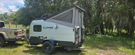 A tan Jeep Gladiator towing a light-colored Clipper pop-up RV rental camper, parked in a green, mossy wooded setting.