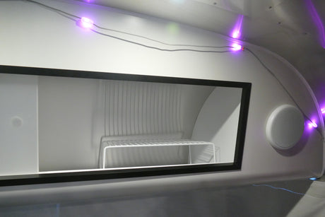 Modern RV rental interior: a clean, white storage unit (likely a refrigerator) with a shelf, illuminated by purple LED lights, and a speaker nearby.