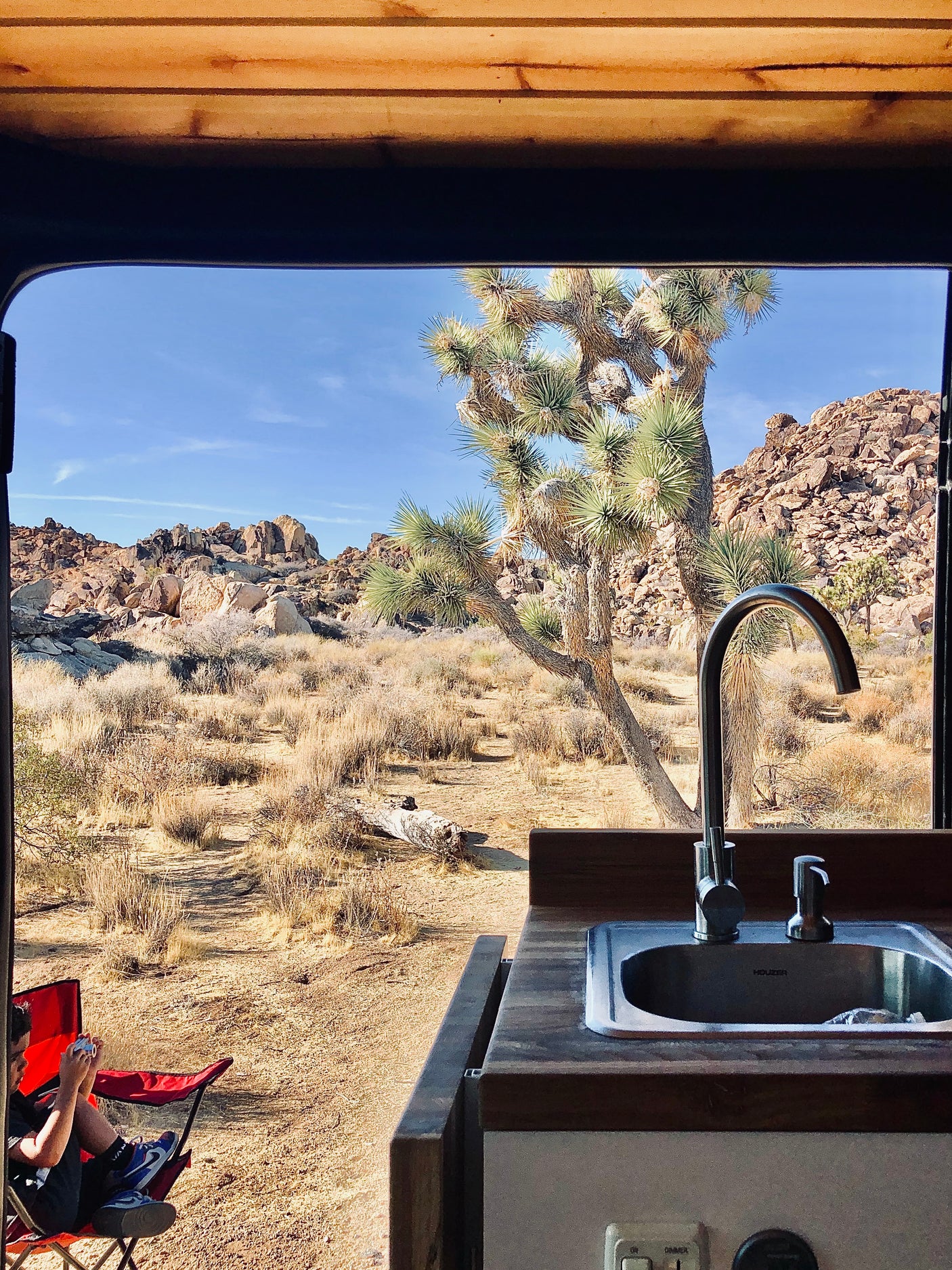 RV rental adventure: Desert landscape with Joshua trees from a camper van's kitchen; person in red chair outside.