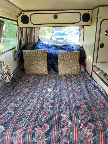 Cozy RV rental interior with a comfortable bed, patterned bedding, and a view of the street through the rear window.