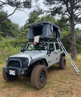 Unique RV rental: Silver Jeep with a rooftop tent and ladder. Bare feet dangle, enjoying an outdoor camping adventure.