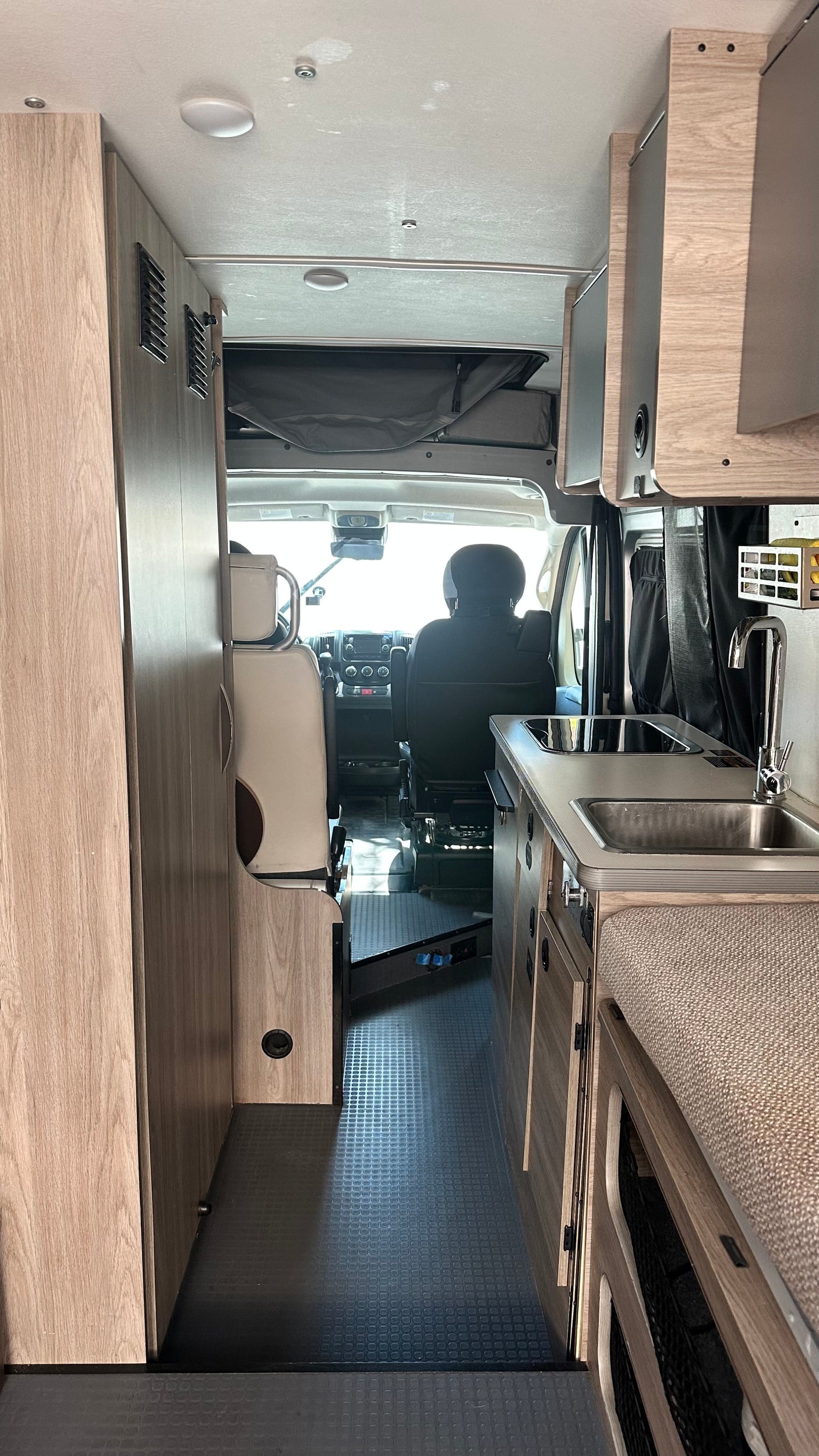 This RV rental interior faces forward, revealing a kitchen with a sink, the driver's cabin, and light wood storage/cabinets.