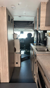This RV rental interior faces forward, revealing a kitchen with a sink, the driver's cabin, and light wood storage/cabinets.