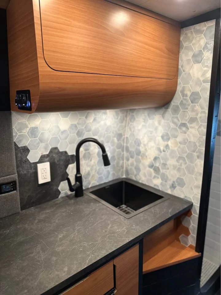 Modern rv rental interior: kitchenette with black sink, dark counter, hexagonal tile backsplash, and warm wood cabinets.