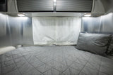 RV rental bedroom with a grey quilted bed, aluminum walls, white fabric curtains, and overhead lights.