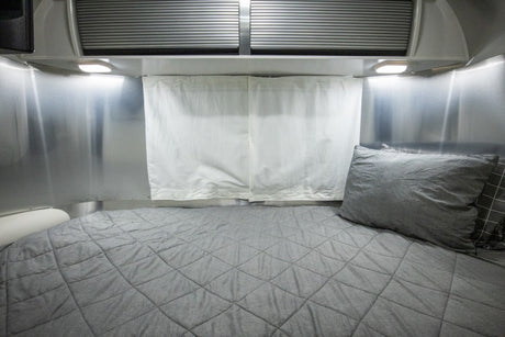 RV rental bedroom with a grey quilted bed, aluminum walls, white fabric curtains, and overhead lights.