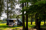 Two friends relax outside their rv rental camper van, parked under shady trees in a bright, grassy field.