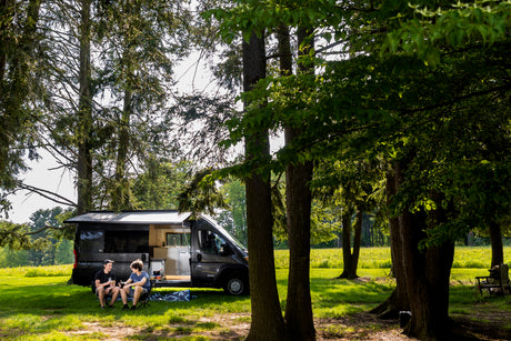 Two friends relax outside their rv rental camper van, parked under shady trees in a bright, grassy field.