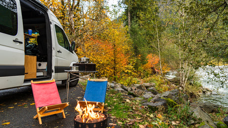 Fall RV rental camping: A camper van beside a vibrant autumn river, with a crackling fire and cozy chairs.
