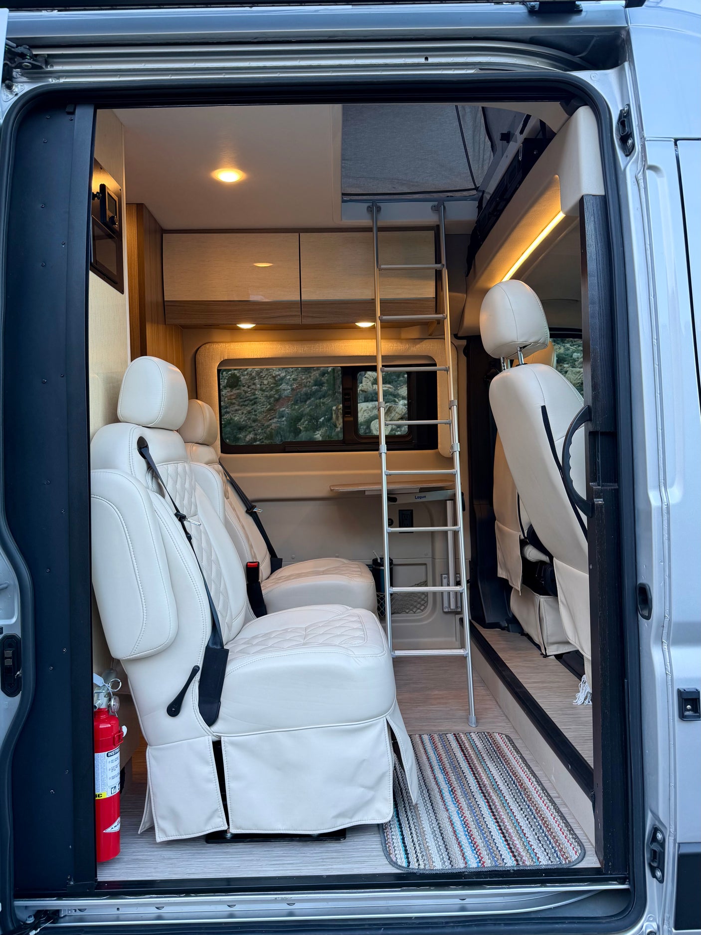 Inside an RV rental: inviting interior with cream seats, an overhead bunk ladder, cabinets, and a scenic view out the window.