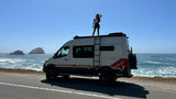 White camper van (RV rental) by the Pacific Ocean. A person stands on the roof, enjoying the coastal vista.