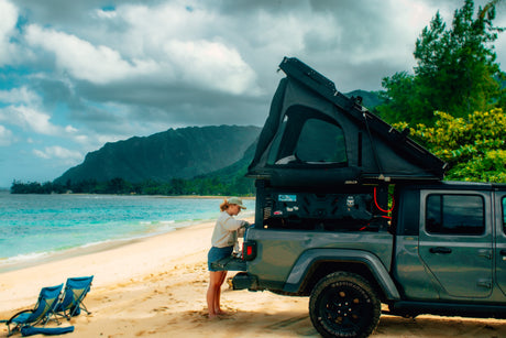 Woman by her truck with a rooftop tent on a stunning beach, mountains in background. Your RV rental adventure awaits!