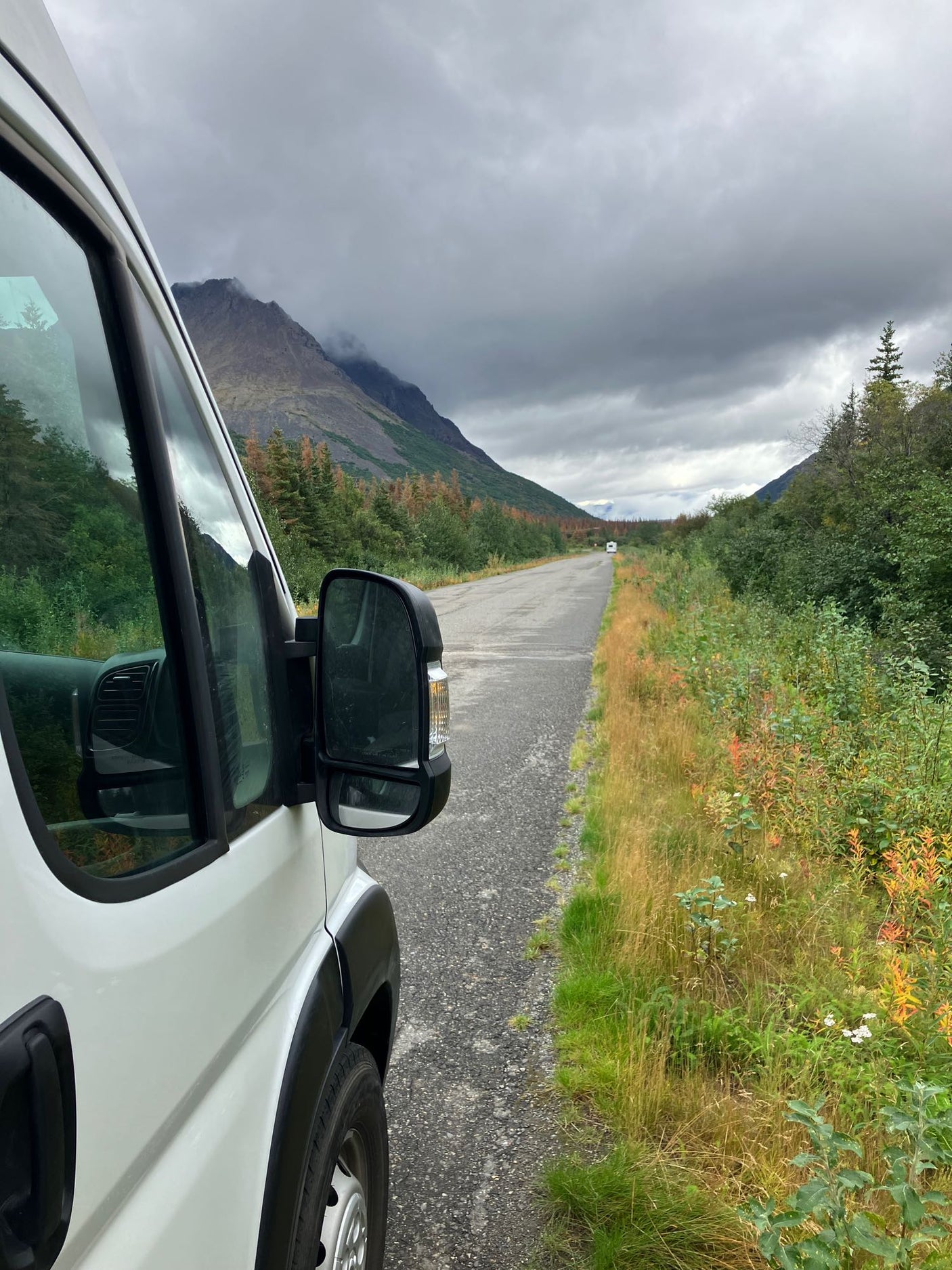 RV rental van on a remote mountain road, leading to another vehicle, framed by cloudy mountains & fall colors.
