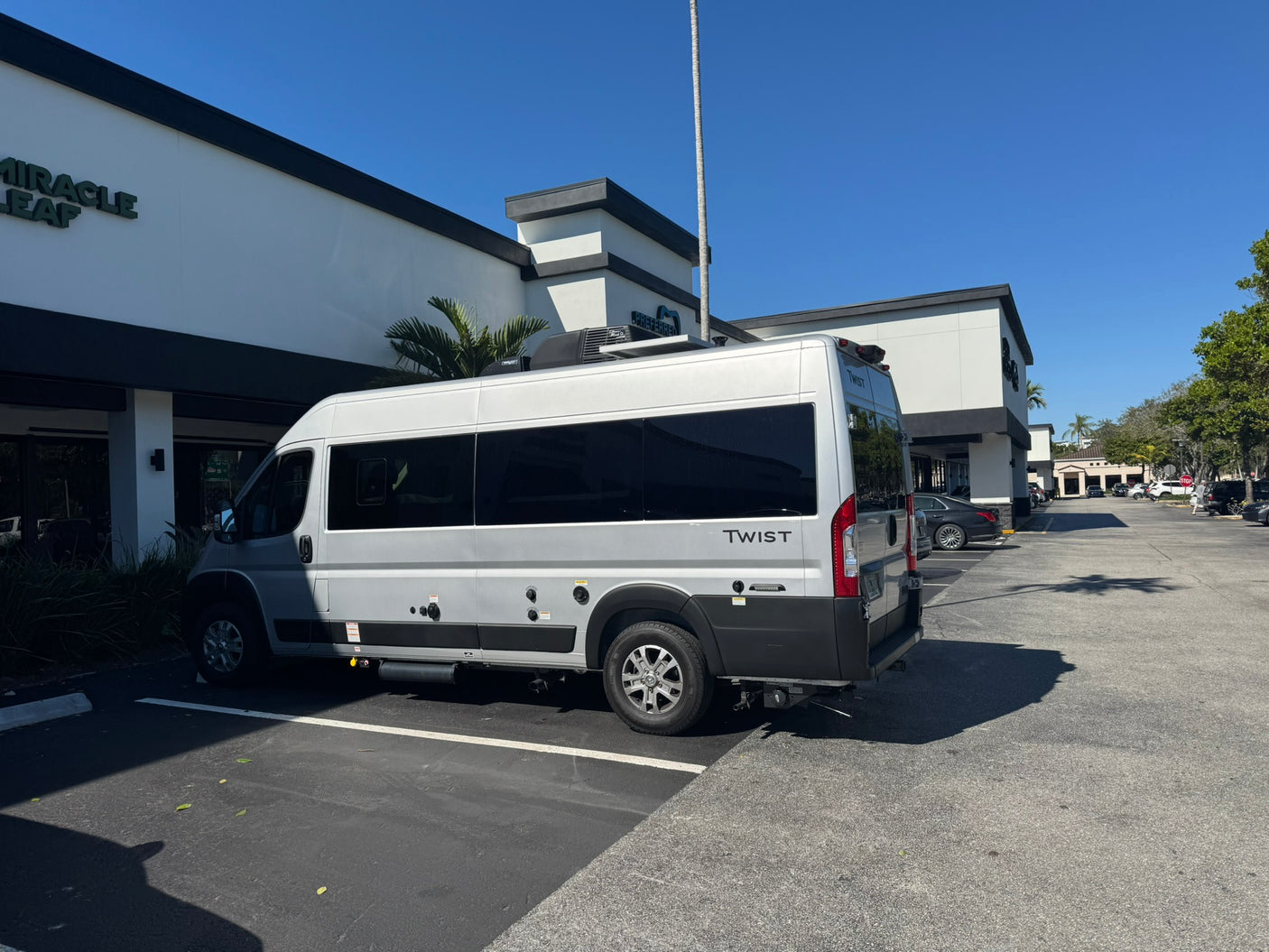 A silver TWIST camper van, an RV rental, is parked in a sunny lot. This Class B motorhome sits before a modern white building.