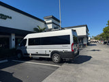 A silver TWIST camper van, an RV rental, is parked in a sunny lot. This Class B motorhome sits before a modern white building.