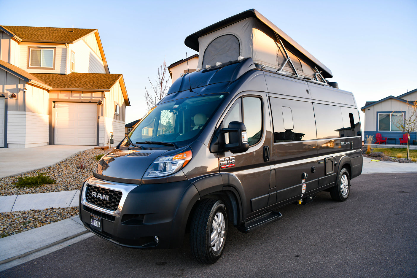 A dark gray Ram ProMaster pop-top RV rental parked on a residential street, with houses in background, bathed in warm sunset light.