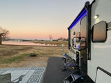 RV rental by a serene lake at sunset. Features a lit travel trailer, outdoor mat, grill, and boats in the tranquil water.