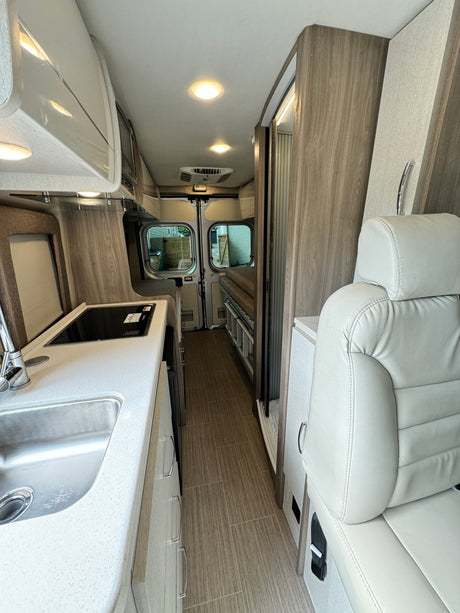 Modern RV rental interior showing kitchenette, light wood cabinetry, and a cozy living area with rear views.