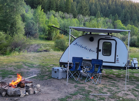 This cozy RV rental camp setup features a Bushwhacker trailer, warm campfire, chairs, cooler, and lush forest.