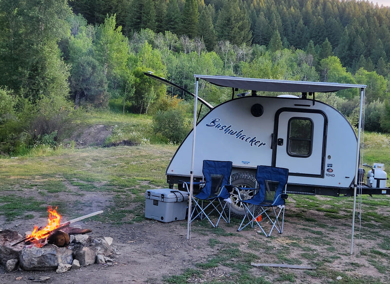 This cozy RV rental camp setup features a Bushwhacker trailer, warm campfire, chairs, cooler, and lush forest. — photo 12