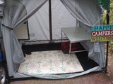 Inside an RV rental's cozy awning: a patterned rug, folding table, and a 'Happy Campers Welcome' sign greet visitors.
