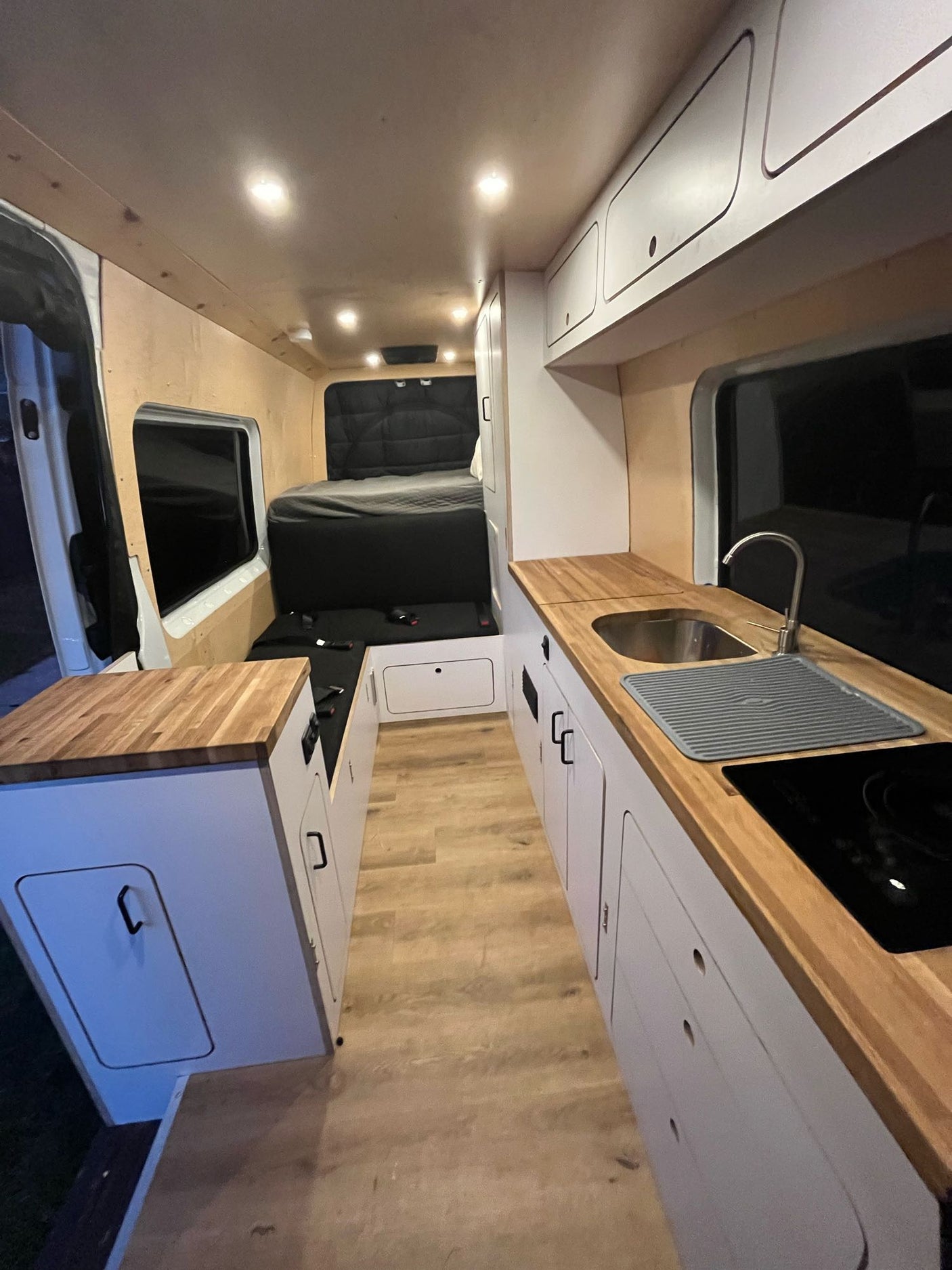 Clean camper van interior: kitchen with wood counters, sink, stovetop; rear bed area; white cabinets. Great for RV rental.