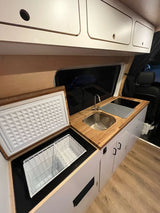RV rental kitchenette with light wood counters, stainless steel sink, cooktop, open top-load fridge, and white cabinets.