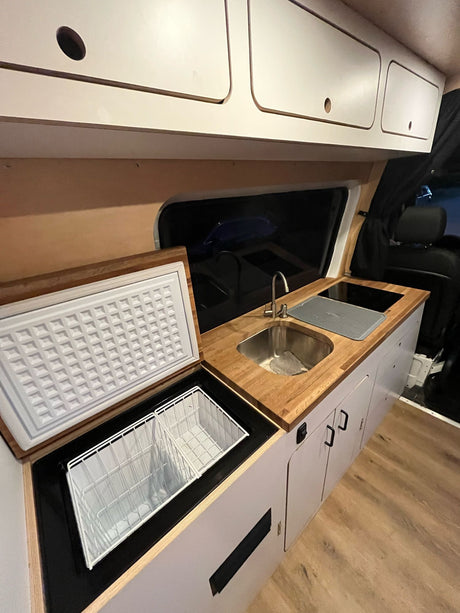 RV rental kitchenette with light wood counters, stainless steel sink, cooktop, open top-load fridge, and white cabinets.