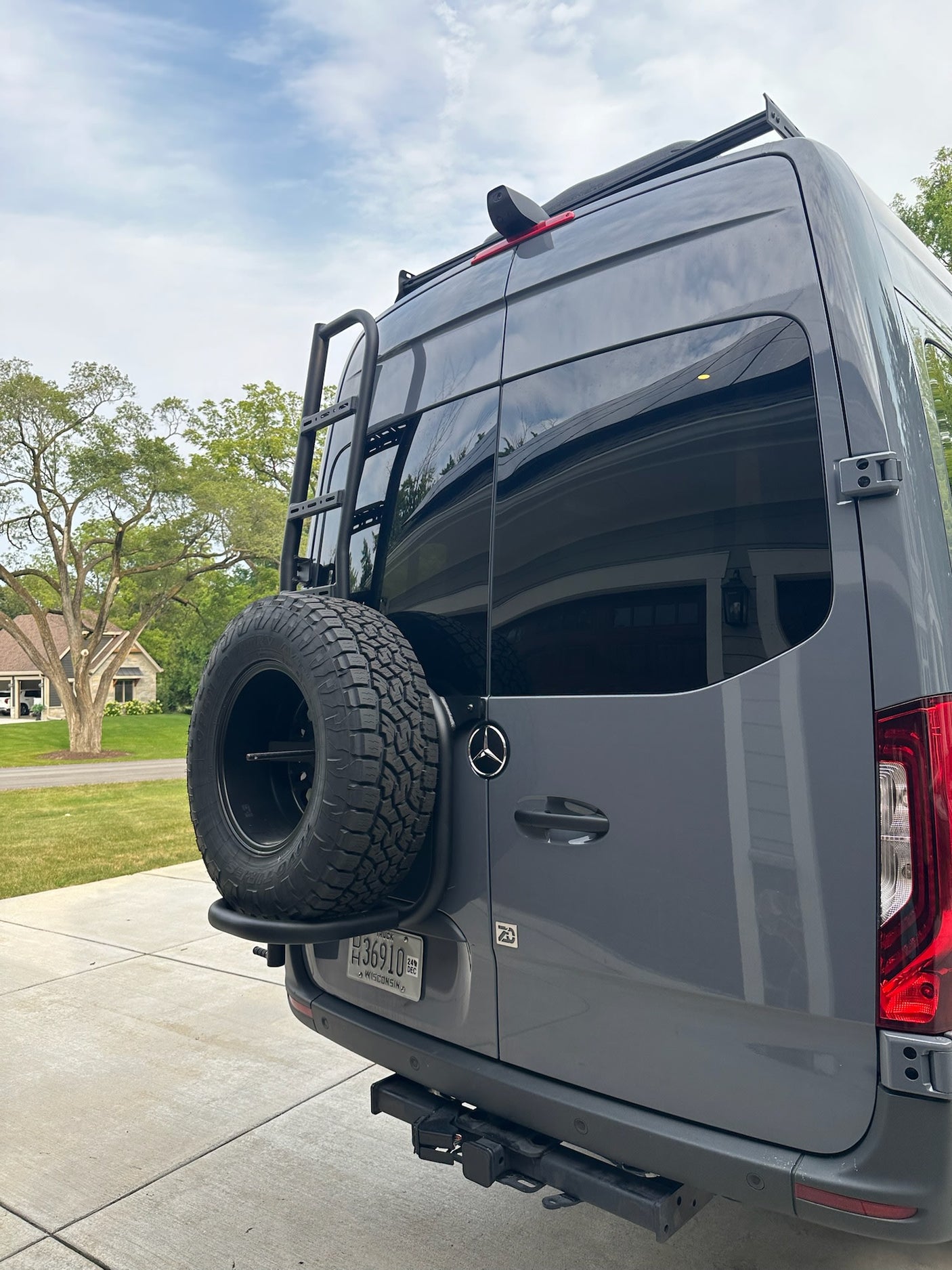 This customized grey Mercedes-Benz Sprinter camper van, available for rv rental, boasts a ladder, roof rack, and spare tire.