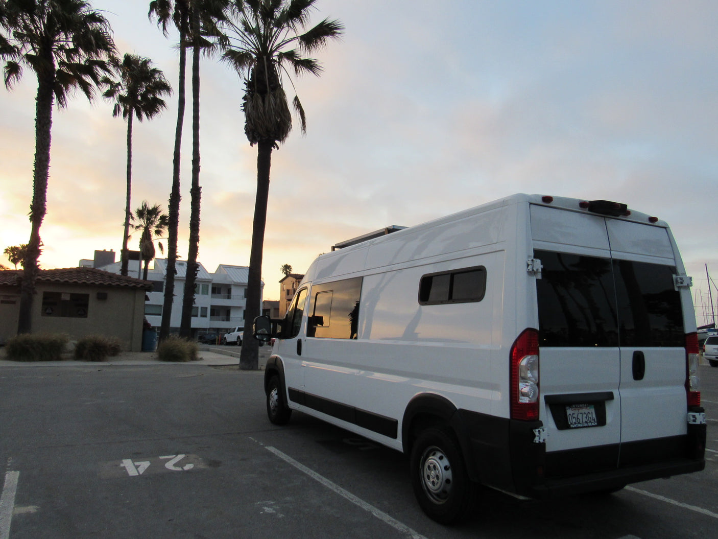 White RV rental camper van parked in a lot at sunset, silhouetted by palm trees and coastal buildings.