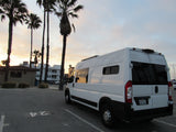 White RV rental camper van parked in a lot at sunset, silhouetted by palm trees and coastal buildings.