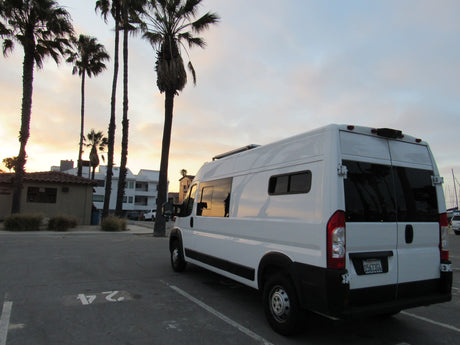 White RV rental camper van parked in a lot at sunset, silhouetted by palm trees and coastal buildings.
