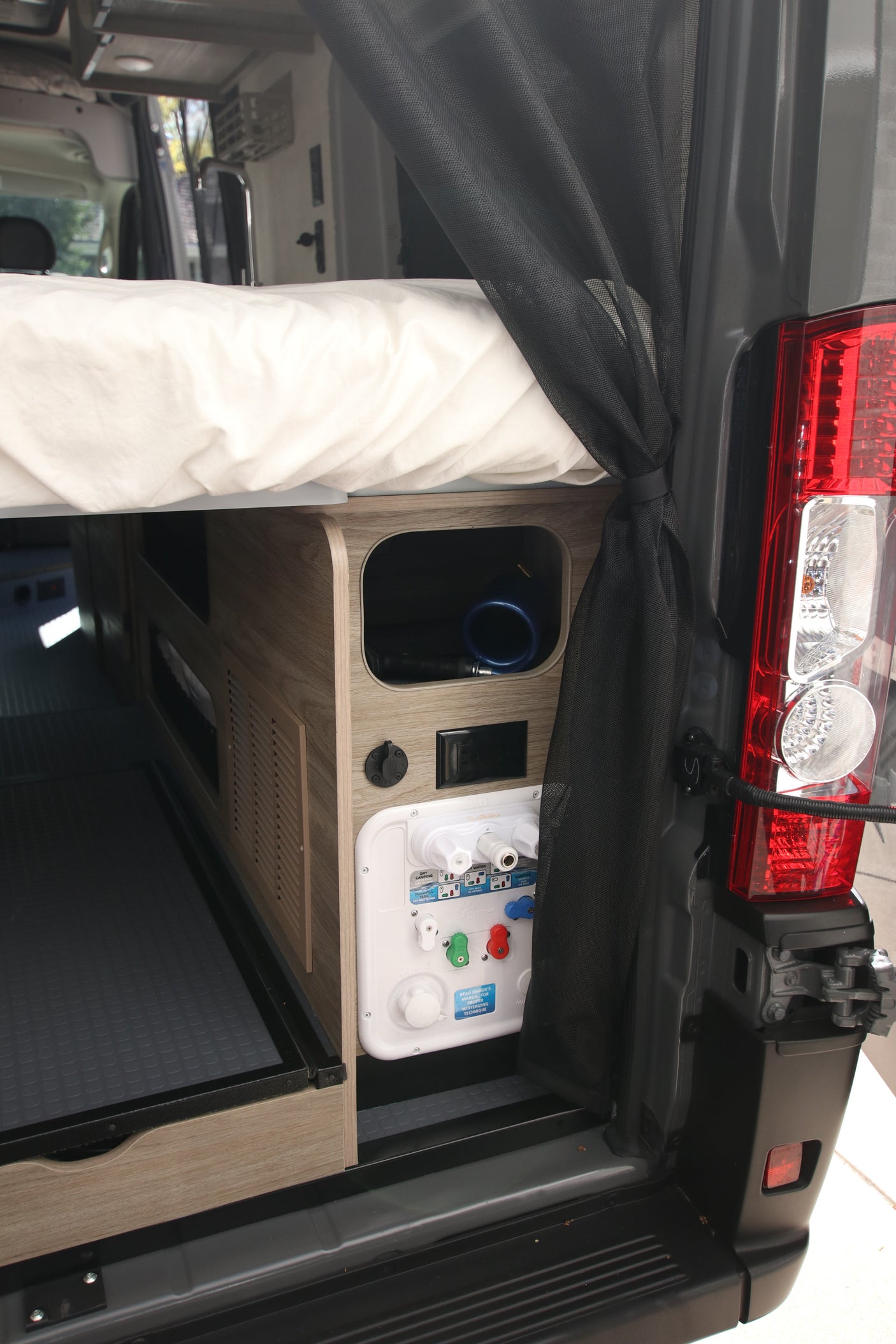 View inside an RV rental: Rear bed and integrated utility panel with water and power connections, plus storage.