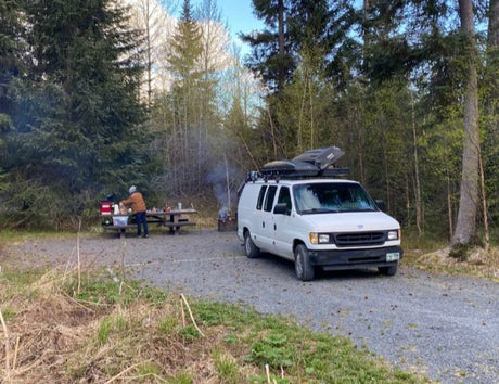 Campsite scene with a white rv rental van, person at picnic table, and campfire in the woods. Perfect for outdoor adventures.