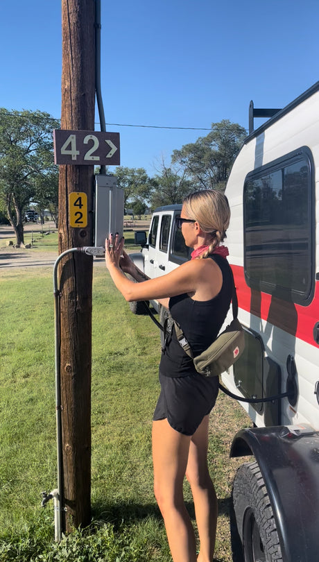 Woman hooking up an RV rental at campsite 42. She's connecting power to a white RV at a sunny campground.