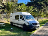 White Mercedes-Benz Sprinter camper van, an RV rental, parked on lush grass near palm trees & picnic tables.