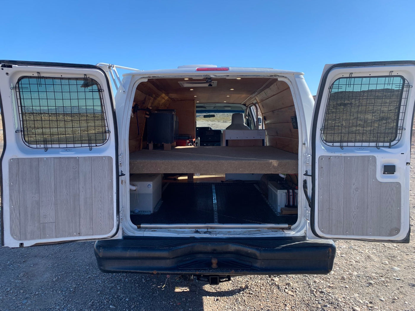  under a clear blue sky. Scenic desert travel.|Off-grid RV rental van with solar panels parked in a rugged Nevada desert landscape near mountains under a clear blue sky.|An RV rental camper van conversion reveals its rear interior with a raised bed platform and organized storage below.|RV rental conversion van: Open side door reveals a compact kitchen with sink — photo 4