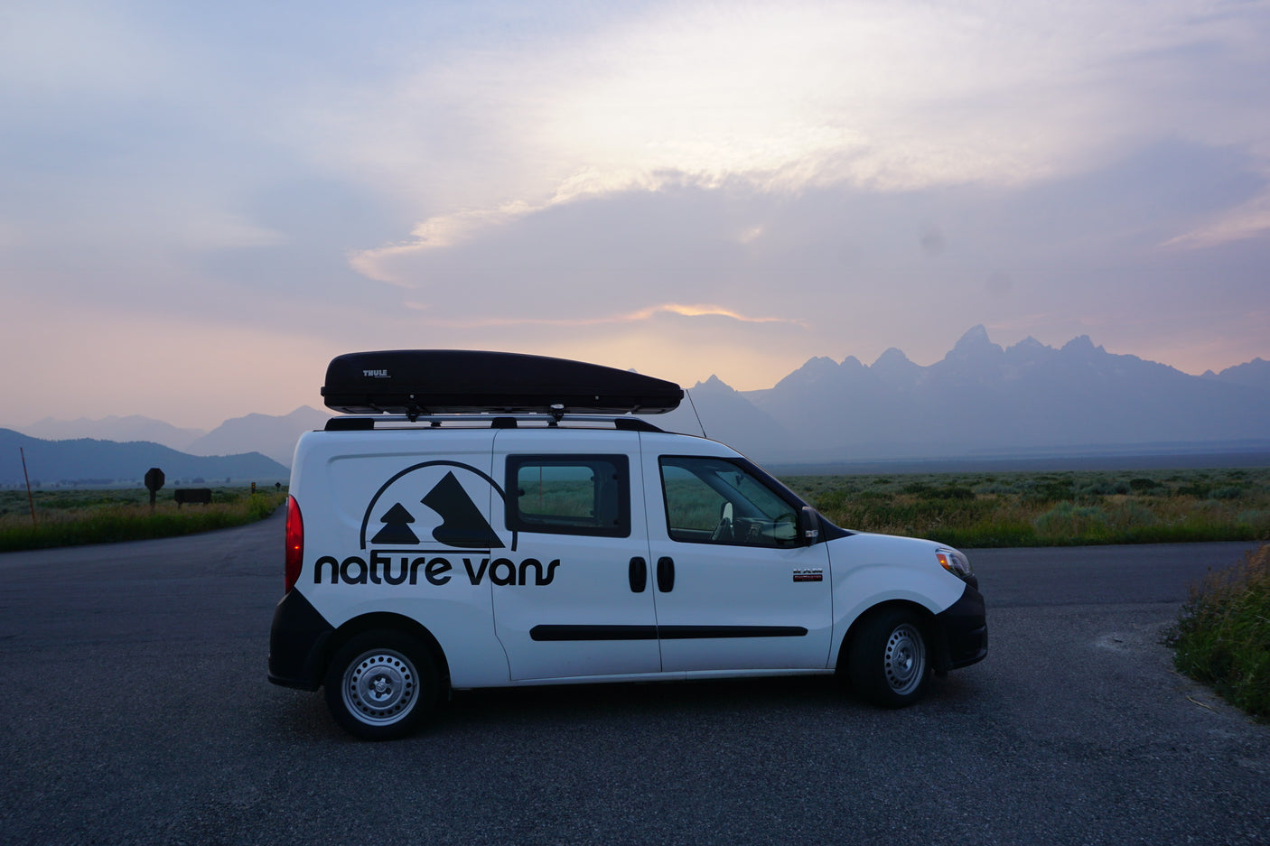 White campervan with roof box, parked on road. Grand Teton mountains and colorful sky. Ideal RV rental for scenic adventures.