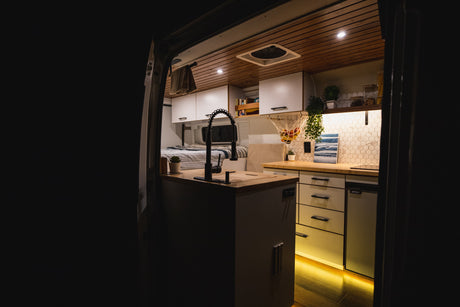 Modern rv rental camper van interior. See the cozy kitchen with warm lights, sleek black faucet, and bed.