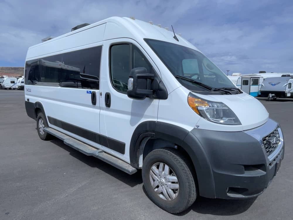 This white RV rental camper van, perfect for road trips, is parked on asphalt with other trailers in the background.