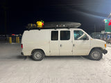 White conversion van with rooftop cargo (tire, jerry cans, cargo box) on snowy ground at night.