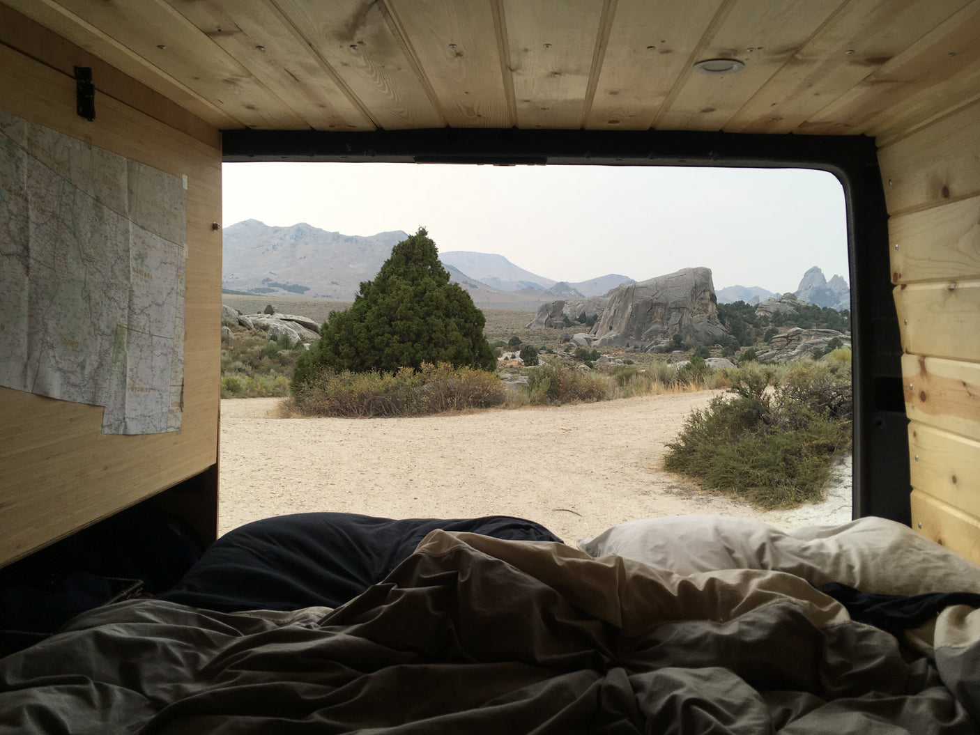 Serene RV rental view from bed: wooden camper interior opens to a desert landscape with mountains & large rocks.