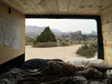 Serene RV rental view from bed: wooden camper interior opens to a desert landscape with mountains & large rocks.