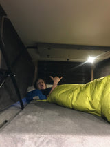 A child makes a shaka sign and shouts in a bright yellow sleeping bag, enjoying a cozy bed setup in an RV rental.